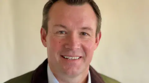 BBC Man with dark brown hair posing for a photo with a cream wall behind him, wearing a pale blue shirt and green jacket with brown collar.