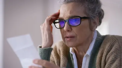 Getty Images Woman in glasses and warm clothes looks at a receipt or bill.