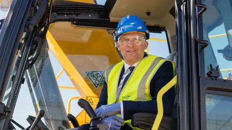 Ministry of Justice James Timpson sitting in the cab of a construction vehicle. He is wearing a blue hard hat and a yellow high-vis jacket over a navy suit. He is wearing clear  protective goggles. 