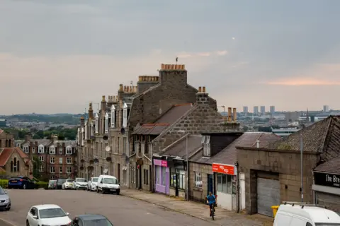 Getty Images An image of a street in Torry, Aberdeen. We see terraced flats and a row of shops with mixed heights. There are hills and high rise flats in the background.