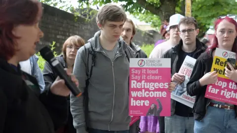 A group of protestors outside the new councils' first meeting, some holding placards. One of the placards reads:
