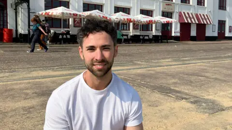 Adam, a young man with dark hair wearing a white t-shirt, sitting in front of a restaurant near the beach in Bournemouth