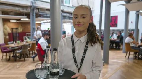 BBC / Gemma Laister Isabel is smiling into the camera while holding a tray with two bottles of water and two glasses on it. She is at work at a cafe with people sat at tables in the background, and is wearing a smart white shirt buttoned up to her neck.