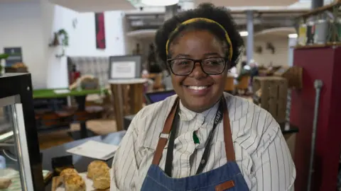 BBC / Gemma Laister Chachomwe smiles into the camera from behind the counter of the cafe she is working at as part of her apprenticeship. She is wearing a blue denim apron over a white striped shirt and has on glasses and a yellow headband.