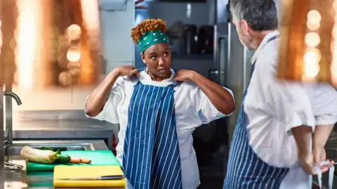 Getty Images A stock photo of a trainee chef adjusting her apron in a kitchen while looking at her older mentor. There are ingredients on the counter in front of her.