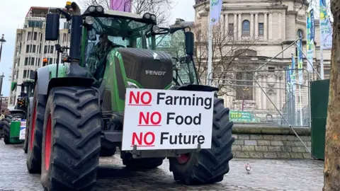 Malcolm Prior/BBC A green tractor is parked on a Westminster pavement under a grey winter sky next to a bare tree and in front of the QEII Centre. On the front of the tractor is a protest placard that reads No Farming, No Food, No Future. Pedestrians pass by.