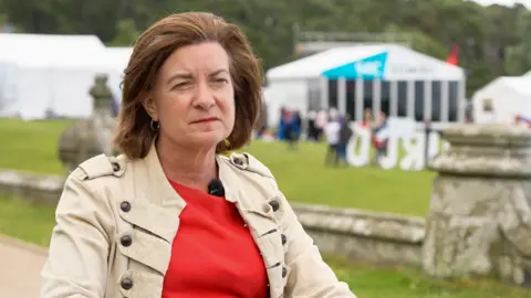 BBC A woman with bobbed dark brown hair, wearing a red blouse and beige jacket and sat with a green field with white tents in it behind her.