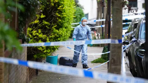 PA Media A person wearing scrubs, hair and face marks, and gloves walks on a street behind police tape