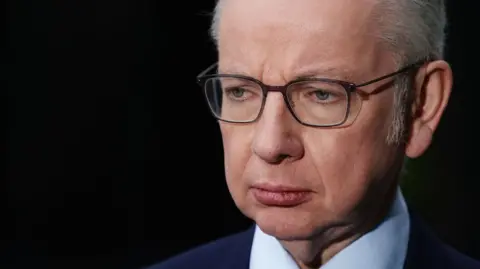 PA images A close-up shot of the former MP Michael Gove, a grey-haired man with glasses. He looks off to his right with a serious expression. He is in a suit and tie and the background is completely black.