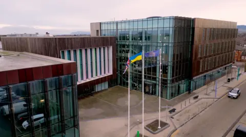 BBC Redcar and Cleveland Council's headquarters photographed from high up. There are three, interlinked square brown buildings. They are clad with the two on either side containing large glass frontages. The middle building, which sits back slightly from the other two, has a large area of wood-effect cladding with windows in the front separated by long, thin white pillars. In front stand three large flagpoles flying the flags of, left to right, the United Kingdom, Ukraine and the council's crest in gold on a blue background.