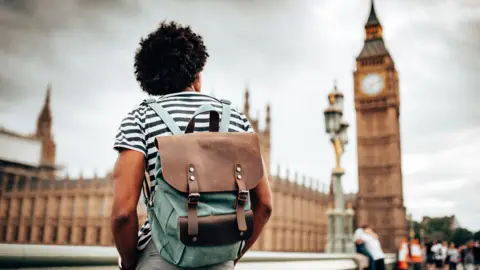 Getty Images A man with dark brown afro hair standing with his back to the camera wearing a black and white horizontal striped t-shirt and a brown and green backpack looking up at the Houses of Parliament