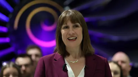 PA Media Rachel Reeves wearing a purple suit, standing in front of a purple spiral logo