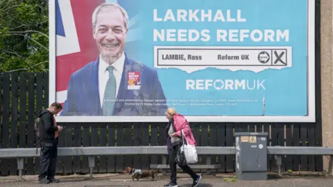 PA Media A large billboard image for reform featuring Nigel farage and a Union flag, two people walk in the foreground.