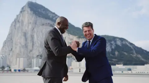 FCDO UK Foreign Secretary David Lammy and Chief Minister of Gibraltar Fabian Picardo clasp hands laughing, as they stand in front of the rock of Gibraltar.