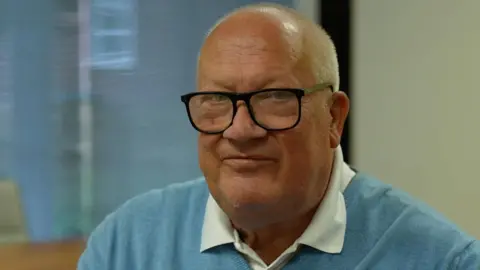 A bald man with large black glasses wearing a blue sweater looks towards the camera