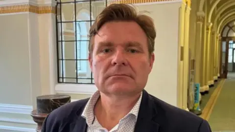 BBC Council leader John Cotton pictured with a blank expression on his face. He has light brown hair which is styled in a quiff. He is wearing a dotted white shirt underneath a blue navy blazer