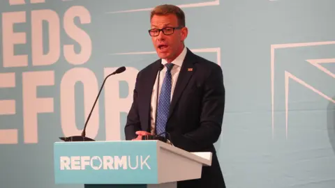 PA Media David Bull gives a speech behind a blue podium, reading 