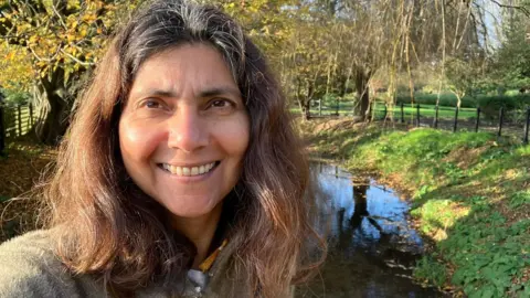 Pallavi Devulapalli Dr Pallavi Devulapalli, former Green Party health spokesperson, standing outdoors near a small stream with autumn trees and a clear blue sky in the background.
