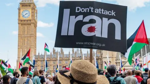 Getty Images A crowd of people face the Houses of Parliament. In the foreground is a person wearing a straw hat and holding a sign that reads: Don't attack Iran