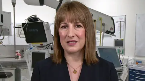 BBC Rachel Reeves wearing a dark suit jacket and gold necklace, looking straight at the camera, with medical equipment seen behind her