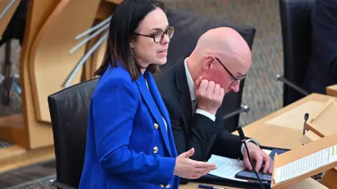Getty Images Kate Forbes is speaking in the Scottish Parliament with John Swinney sitting next to her. Forbes has dark shoulder-length hair and is wearing black framed glasses and a blue jacket over a white top. Swinney is looking down at his phone. He is a bald man with glasses and is waring a black jacket over a white shirt