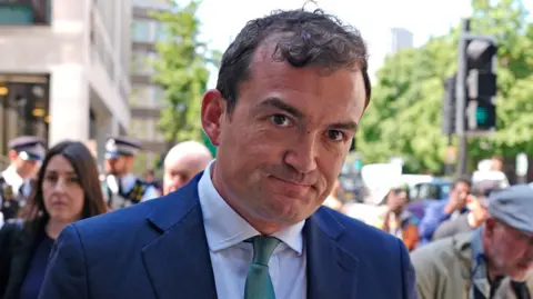 PA Media Patrick Spencer leaves a court, dressed in a blue suit and green tie. He is smiling at the cameras. In front of him is a police officer, dressed in uniform. 