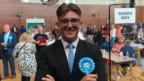 BBC George Finch pictured smiling while crossing his arms. He is wearing wide round black framed glasses and is in a black suit jacket with a white shirt underneath and a blue tie.