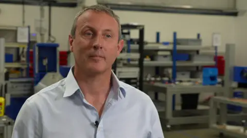 Mick Crosthwaite stands in a factory wearing a grey shirt.