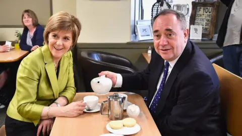Getty Images A man with thinning dark hair and a woman with short fair hair sit opposite each other at a table in a cafe. The man, on the right, is holding a pot of tea above the woman's cup. He is wearing a dark suit, she is wearing a light green jacket. They are both smiling. There are biscuits, a coffee pot and a milk pourer on the table.