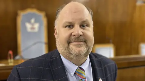 Jim Robbins wearing a multi coloured tie and cheque navy suit jacket standing in the council chambers