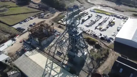A pit shaft metal gear above the surface with a mass of metal girders and a car park behind along with a number of industrial buildings.