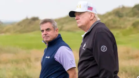 PA Media Two men walk along the fairways of a golf course. President Trump is wearing a black half zip windcheater and a white baseball cap, with USA in gold letters. It also has a saltire on the side. Paul McGinley has brown/ grey hair combed in a side shed . He is wearing a purple polo shirt and a sleeveless blue half zip. Both men are looking off to the left.