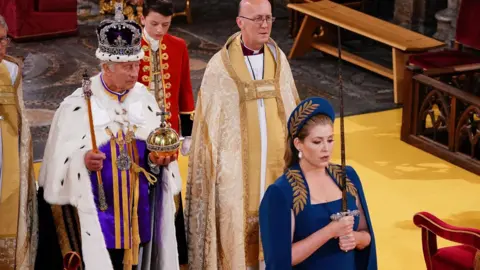 PA Media A newly-crowned King Charles III stands holding an orb and sceptre, while Penny Mordaunt, wearing a blue dress and hat, stands in front of him holding a sword with both hands. The sword is held upright in front of her face.  