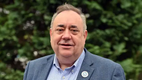 Getty Images A man with grey thinning hair stands in front of greenery. He is looking at the camera and is visible from the chest up. He is wearing a dark blue jacket over a blue and white chequered shirt. An Alba badge with a saltire on it is pinned to his jacket.