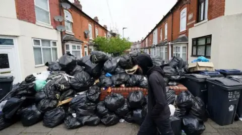 PA Media Man in a black hood walking past high pile of black binbags.