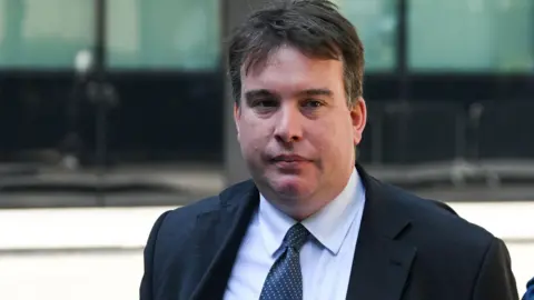 Reuters A man in a suit is photographed. He has brown hair and is wearing a blue tie. The arm of another person can be seen on the right of the image.