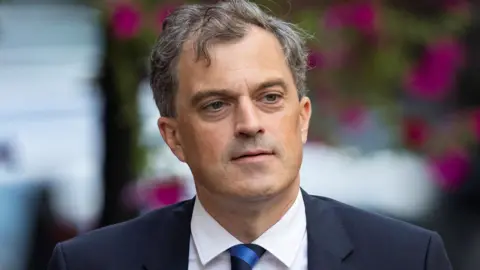 Getty Images Julian Smith is outside. Flowers are behind him. He's wearing a navy suit.