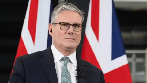EPA Sir Keir Starmer, dressed in a dark suit and light green tie, stands at a podium speaking. Behind him are two large Union Jack flags. His face is blurred in the image.
