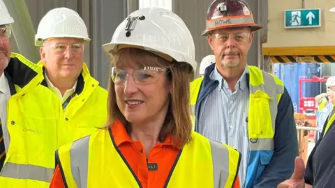 Seb Noble/BBC Chancellor Rachel Reeves is wearing an orange top and yellow high vis jacket and a white hard hat. Several men who are wearing similar protective clothing are standing behind her.