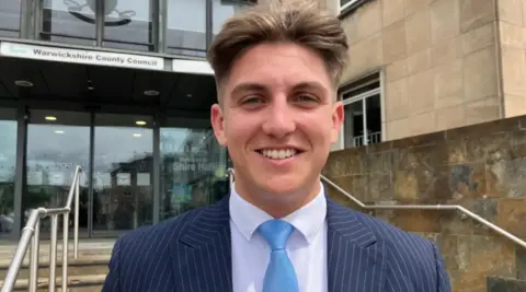 BBC A young man with short light brown hair is wearing a blue pinstriped suit, a white shirt and pale blue tie. In the background are steps leading up to a large glass building with a sign above double doors that reads, 