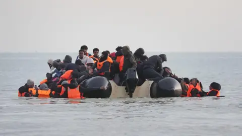 PA Media A small, crowded inflatable boat filled with people in bright orange life jackets departs from the beach at Gravelines, France. Several individuals are still in the shallow water, attempting to climb aboard.