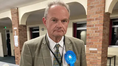 LDRS A man with short grey hair, a grey jacket, green tie and a blue Conservative Party rosette, stands in a hall with brick pillars and a white wall with windows behind him.