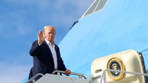 Reuters President Trump, in dark blue suit and open-necked white shirt, waves as he boards his plane. The sky is blue.
