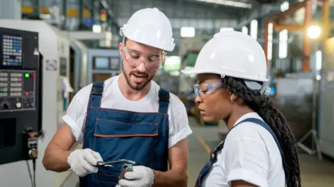 Getty Images A male and female engineer both wearing white hard hats discussing a piece of machinery