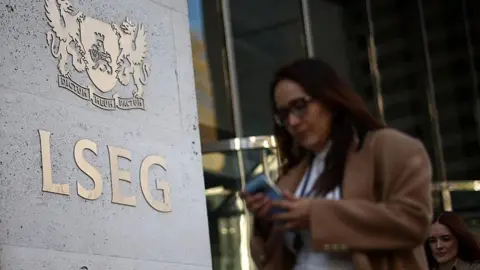 Getty Images A woman wearing a brown coat walks by the London Stock Exchange in London, she's on her phone and is wearing glasses