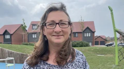 Lindsey is wearing rectangular glasses and has shoulder length hair. She is standing in a playground in front of some new houses, with a roundabout and a goal net either side of her in the background
