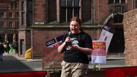 Councillor Grace Lewis A woman in a black T-shirt with a white graphic on the front and brown trousers is talking into a microphone while looking at her mobile phone. Behind her are placards about university cuts