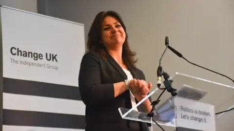 Getty Images Heidi Allen, a woman with shoulder length black hair, dressed in a black suit, smiles and clasps her hands on a Change UK party podium. The slogan on the podium says