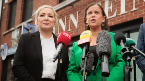 PA Media Michelle O'Neill, a woman with shoulder-length blonde hair, standing beside Mary Lou McDonald during a Sinn Féin press conference. McDonalds has short, dark hair andis wearing a black top and a bright green blazer. O'Neill is weaing a white blouse and black suit jacket.