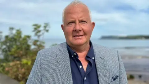 Shane Laverty is wearing a grey suit jacket and navy shirt.
He is standing near a body of water and a tree is visible beisde him.
